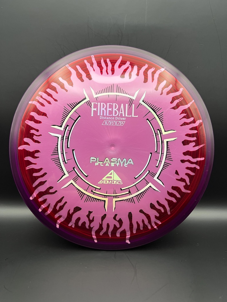 May include: A pink and red Fireball distance driver disc with the text "Fireball" and "Plasma Plastic" printed on it. The disc has a unique design with a central circular pattern and flame-like edges. The disc is made by Axiom Discs.