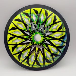 May include: A round disc golf driver with a black rim and a yellow, green, and blue marbled center. The graphic features a black geometric starburst pattern, silver text reading TRAIL, JAMES CONRAD | 2021 WORLD CHAMPION, and flight numbers 10 | 5 | -1 | 1.