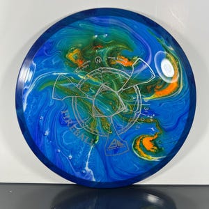 May include: A blue, green, and orange disc golf disc with the word "Fireball" and other text. The disc has a swirled pattern of colors and a silver graphic in the center. The disc is a distance driver.