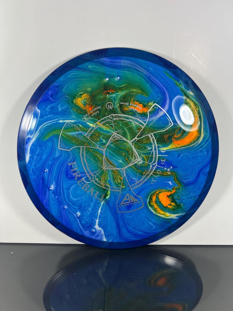 May include: A blue, green, and orange disc golf disc with the word "Fireball" and other text. The disc has a swirled pattern of colors and a silver graphic in the center. The disc is a distance driver.
