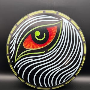 May include: A black disc golf disc featuring a stylized eye design. The eye is red with a green pupil, surrounded by white and black wavy lines. The disc has a yellow and green border.