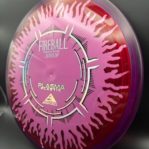 May include: A disc golf disc in shades of purple and red, featuring the word "FIREBALL" and "PLASMA PLASTIC". The disc has a flame design around the edge and the text "Distance Driver" is also printed on the disc.