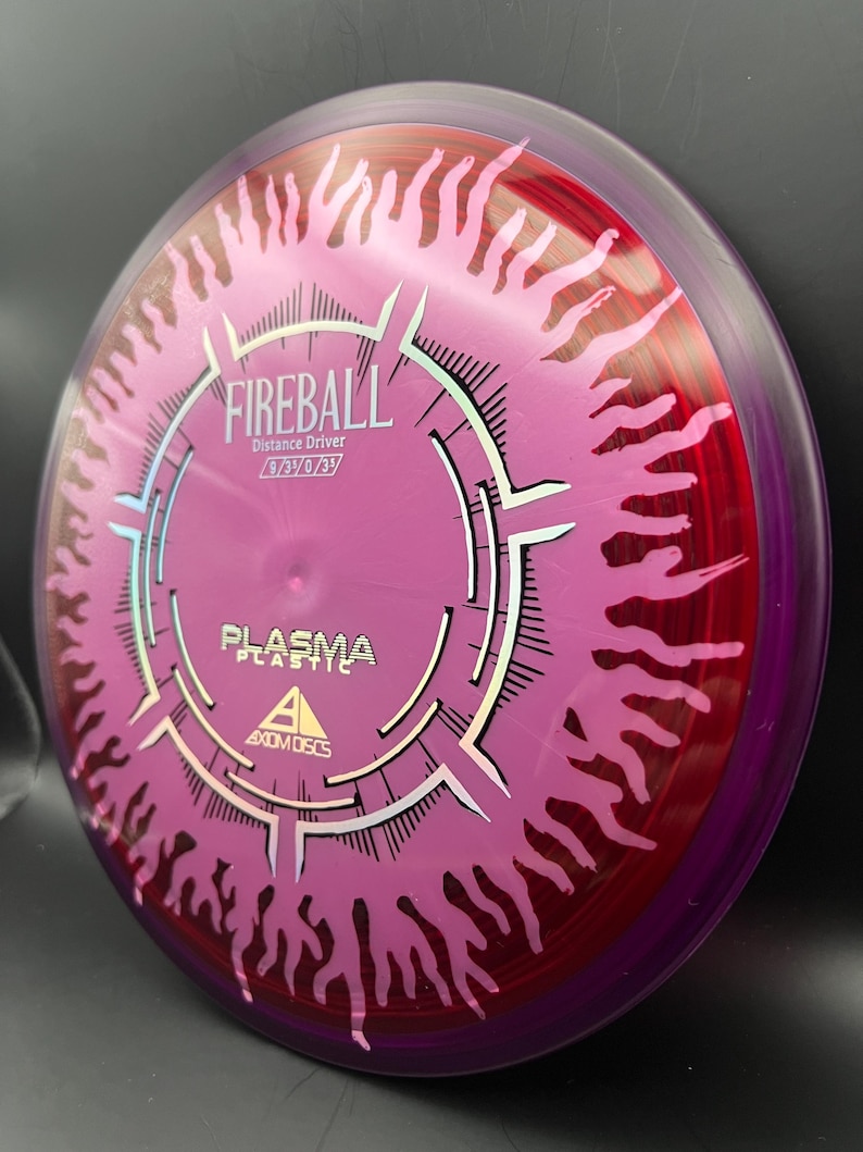 May include: A disc golf disc in shades of purple and red, featuring the word "FIREBALL" and "PLASMA PLASTIC". The disc has a flame design around the edge and the text "Distance Driver" is also printed on the disc.