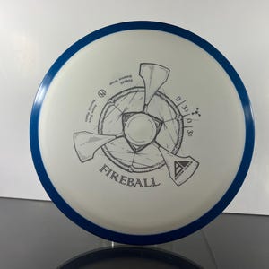 May include: A white disc golf disc with a blue rim. The disc features a gray graphic design of a propeller-like shape with the word "FIREBALL" and the Axiom Discs logo. The disc has the numbers 9/3/0/3.5 printed on it.