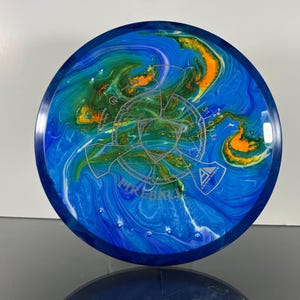 May include: A blue and white disc golf disc with a swirling pattern of blue, green, and orange. The disc has the word "FIREBALL" and other text printed on it. The disc has a dark blue rim and a silver graphic in the center.