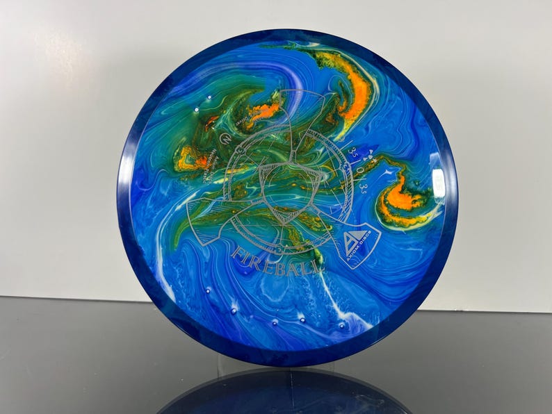 May include: A blue and white disc golf disc with a swirling pattern of blue, green, and orange. The disc has the word "FIREBALL" and other text printed on it. The disc has a dark blue rim and a silver graphic in the center.