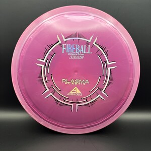 May include: A pink and purple disc golf disc featuring the word "FIREBALL" in white. The disc also has the text "Distance Driver" and "PLASMA PLASTIC" printed on it. A disc golf distance driver.