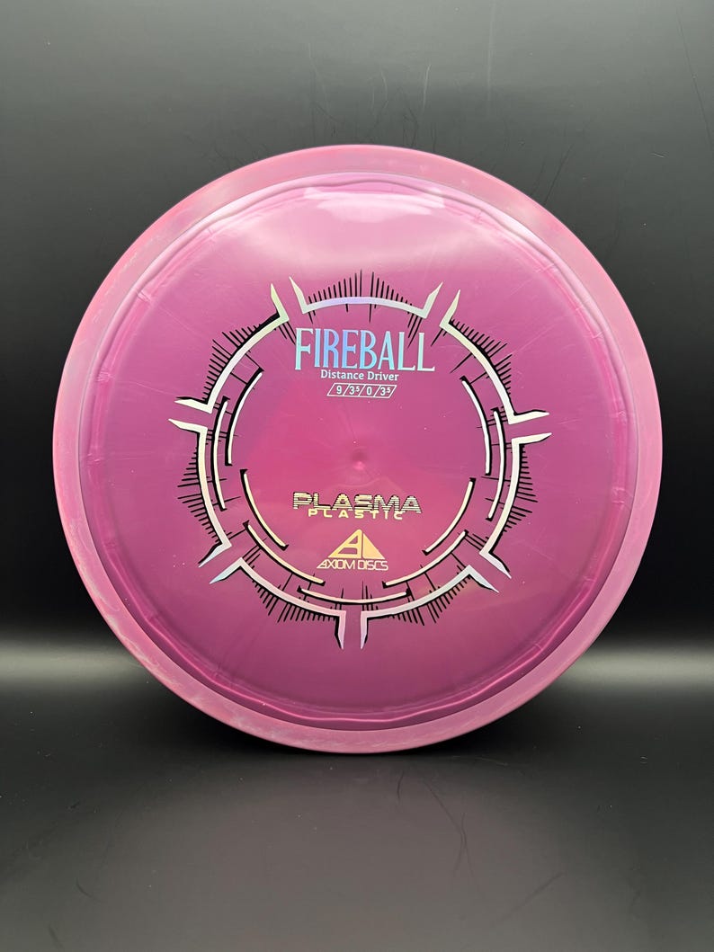 May include: A pink and purple disc golf disc featuring the word "FIREBALL" in white. The disc also has the text "Distance Driver" and "PLASMA PLASTIC" printed on it. A disc golf distance driver.