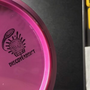 May include: A pink disc golf disc with a black logo that says "DISCDYEADDICT". The disc has a small, raised pink dot and the text "1/19". The disc is on a black surface.