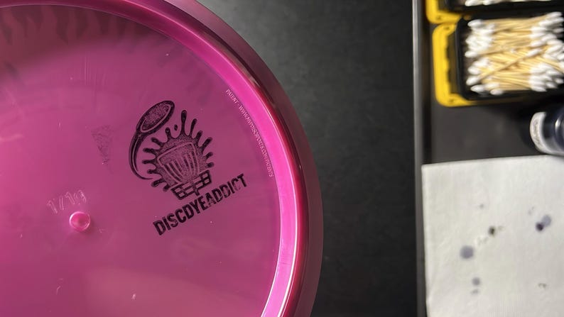 May include: A pink disc golf disc with a black logo that says "DISCDYEADDICT". The disc has a small, raised pink dot and the text "1/19". The disc is on a black surface.