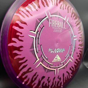 May include: Axiom Discs Fireball distance driver disc in plasma plastic. The disc is primarily pink with a red and purple rim. The disc has a flame design and the word "Fireball" is printed on the disc.