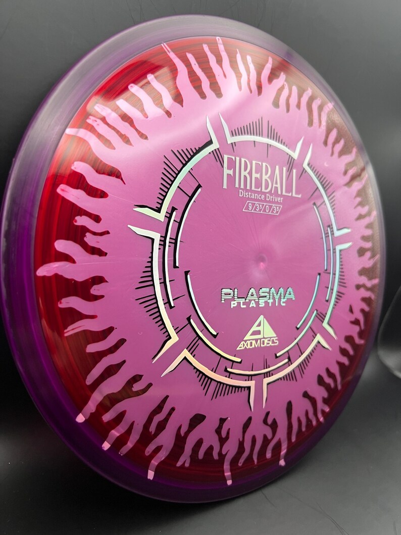 May include: Axiom Discs Fireball distance driver disc in plasma plastic. The disc is primarily pink with a red and purple rim. The disc has a flame design and the word "Fireball" is printed on the disc.