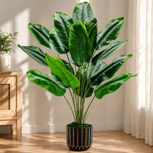 May include: A tall artificial plant with large, vibrant green, oval-shaped leaves extending outward from a central stem. The plant is housed in a cylindrical black pot featuring a vertical gold-toned geometric pattern around the base.