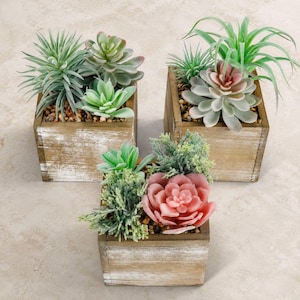 Puede incluir: Tres maceteros cuadrados de madera con un acabado blanco desgastado, cada uno con una variedad de suculentas artificiales. Las plantas presentan tonos verdes, salvia y rosa empolvado, con pequeñas piedras visibles en la base de cada recipiente.