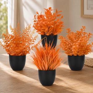 May include: Four artificial plants with vibrant orange foliage, each potted in a small, matte black container. The plants feature varied leaf shapes, including fern-like fronds, rounded leaves, and spiky grass-like blades, arranged in a group.