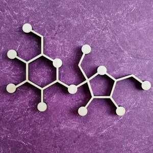 May include: A wooden model of a chemical structure with six rings and multiple branches. The model is made of light brown wood and is set against a purple background.