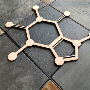 Op de afbeelding: Een houten uitsnijding van een chemische structuur, die lijkt op een hexagon met extra ringen en takken, op een grijs betegeld oppervlak.