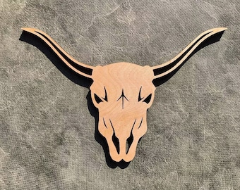 May include: A wooden cutout of a bull skull with a black outline. The skull has a simple arrow design in the center.