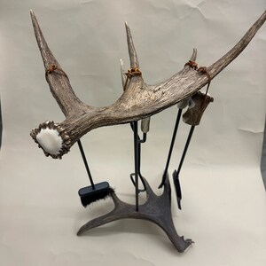 May include: A decorative fireplace tool set featuring a large antler as the main element. The antler is a light brown color with a white tip and is mounted on a black metal stand. The set includes a brush and other tools.
