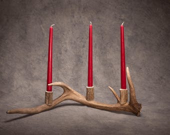 May include: A rustic candle holder crafted from a deer antler, holding three tall red candles. The antler is a natural brown color, and the candles are evenly spaced. A unique home decor item.