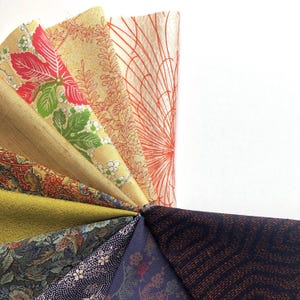 May include: A collection of eight fabric swatches featuring a variety of patterns and colours, including red, green, blue, yellow, and black. The fabrics are arranged in a fan shape, with the tips of the swatches pointing towards the right side of the image.