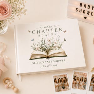 May include: A white hardcover guest book featuring the text A NEW CHAPTER BEGINS, an illustration of an open book with wildflowers and butterflies, and the personalized text OLIVIA'S BABY SHOWER JULY 17TH, 2026, displayed with photos.