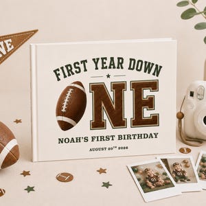 May include: A white hardcover photo book with the text "FIRST YEAR DOWN ONE" and "NOAH'S FIRST BIRTHDAY AUGUST 20TH 2026" in dark green and brown. The cover features a brown football graphic. A small pennant, a football, and three photos are nearby.