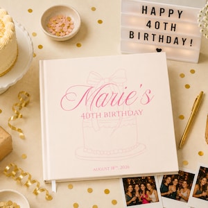 Peut inclure: Un album photo carré à couverture crème avec le texte rose Marie's 40TH BIRTHDAY et AUGUST 18TH, 2026, ainsi qu'un dessin au trait d'un gâteau et d'un nœud. L'album est entouré d'un gâteau d'anniversaire, d'une boîte lumineuse et de photos.