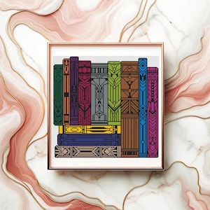 May include: A framed cross-stitch artwork featuring a stack of colorful books. The books have intricate geometric designs in shades of green, purple, blue, orange, and yellow. The artwork is displayed in a gold-colored frame.