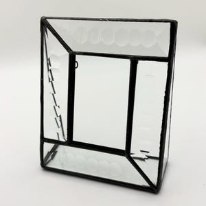 May include: Clear glass terrarium with a geometric design. The glass panels are held together by black metal framing. The top and side panels feature a pattern of small, raised circles. The terrarium is empty, ready for plants or other decorative items.