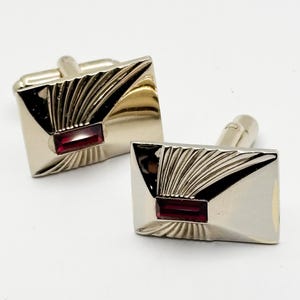 May include: A pair of silver-toned cufflinks with a rectangular shape. Each cufflink features a red rectangular stone in the center and a fan-like design radiating from the top. The cufflinks are set against a white background.