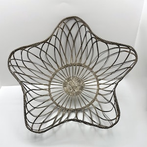 May include: A star-shaped wire basket with a woven centre and a twisted rope-like border. The basket is made of a dark, metallic material and is empty. The design is open and airy, suitable for displaying fruit or other items.