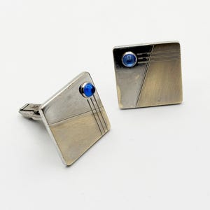 May include: A pair of vintage silver cufflinks with a geometric design. Each cufflink features a square shape with a diagonal split, a blue gemstone, and three parallel lines. The cufflinks have a brushed metal finish.