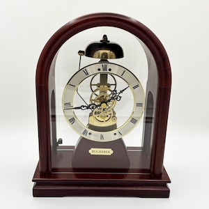 May include: A Bucherer mantel clock featuring a dark wood arched frame with a glass front and back. The clock has a circular white face with black Roman numerals, exposed gold-tone mechanical gears, and a gold-tone bell at the top center.