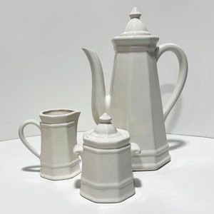 May include: A white ceramic tea set featuring a tall, angular teapot, a small creamer pitcher, and a lidded sugar bowl. The set has a classic, geometric design and a glossy finish, perfect for serving tea or coffee.