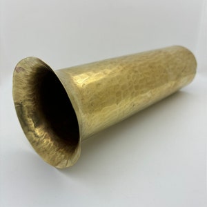 May include: A hammered brass vase with a flared rim. The cylindrical body has a textured surface and a warm, golden hue. The vase is designed to hold flowers or serve as a decorative piece. The vase is approximately 10 inches long.