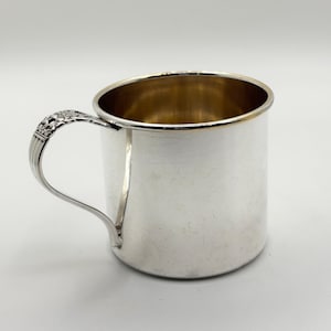 May include: A silver-colored mug with a handle. The handle has a decorative design. The inside of the mug has a gold color. The mug is on a white background.