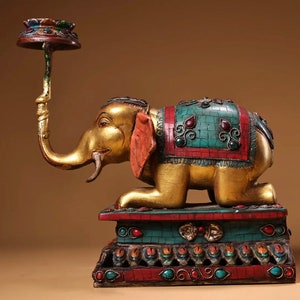 May include: A gold-painted wooden elephant statue with a colorful jeweled saddle and a decorative candle holder on its trunk. The elephant is kneeling on a multi-colored base with intricate details.
