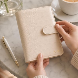 May include: A cream-colored, rectangular personal planner with a textured crocodile-skin pattern. The cover features a magnetic snap closure and the gold-embossed initials A.M. in the bottom right corner. The planner is held in a vertical orientation.