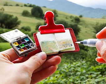 Könnte beinhalten: Ein kleiner roter Ordner mit einem Clip enthält ein Miniatur-Aquarell-Skizzenbuch. Eine silberne und schwarze Aquarellpalette ist ebenfalls abgebildet. Das Skizzenbuch ist auf einer Seite mit einer Aquarellmalerei einer Landschaft geöffnet.