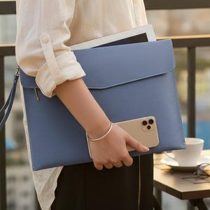 May include: A light blue leather laptop sleeve with a matching wrist strap. The sleeve is held by a person wearing a white button-down shirt and black pants. A smartphone is visible, along with a tablet inside the sleeve.