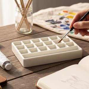 May include: A white ceramic watercolor palette with multiple compartments sits on a wooden table. A hand holds a paintbrush over the palette. Paint tubes, paintbrushes in a glass jar, and a sketchbook are also on the table, suggesting an art studio setting.