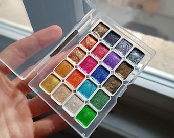 May include: A white plastic palette with 18 round paint pans. The pans are filled with metallic watercolor paints in various colors, including gold, silver, green, blue, red, and purple.