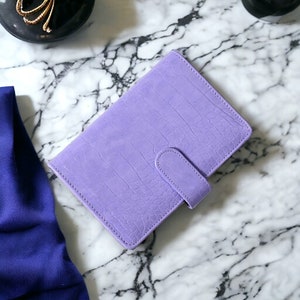 May include: A lavender-coloured, faux-leather planner with a crocodile skin texture. The planner has a tab closure and is closed. The planner is on a marble surface.