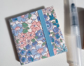 May include: A small, floral patterned notebook with a blue ribbon closure. The notebook is made of fabric and has a white background with pink and blue flowers.