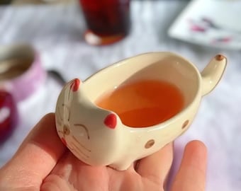 May include: A white ceramic cat-shaped dish with red accents, holding a light orange liquid. The dish is being held in a hand.