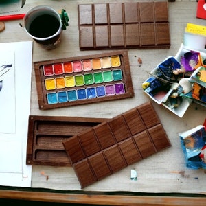 May include: A wooden watercolor palette with a rainbow of paint colors in individual pans. The palette is open and the lid is separate. The lid has a rectangular shape with a grid pattern.