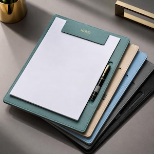 Può includere: Una pila di cartelle portablocco in pelle di vari colori: verde acqua, beige, azzurro, grigio e nero. Ogni cartella contiene un foglio di carta bianco e ha un portapenne. La cartella verde acqua ha la scritta "HOTEL" in rilievo dorato.