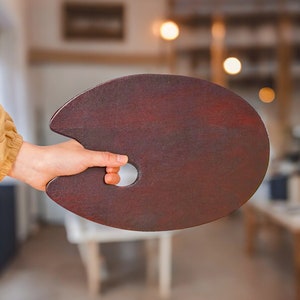 Könnte beinhalten: Eine dunkelbraune Holzpalette für Künstler mit einem Daumenloch. Die Palette wird in einer Hand gehalten.