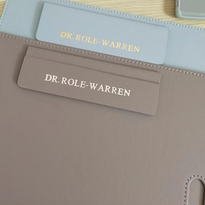 Può includere: Un organizer da scrivania in similpelle grigio chiaro e azzurro con la scritta "DR. ROLE-WARREN" stampata in oro. L'organizer ha una tasca e un bordo cucito.